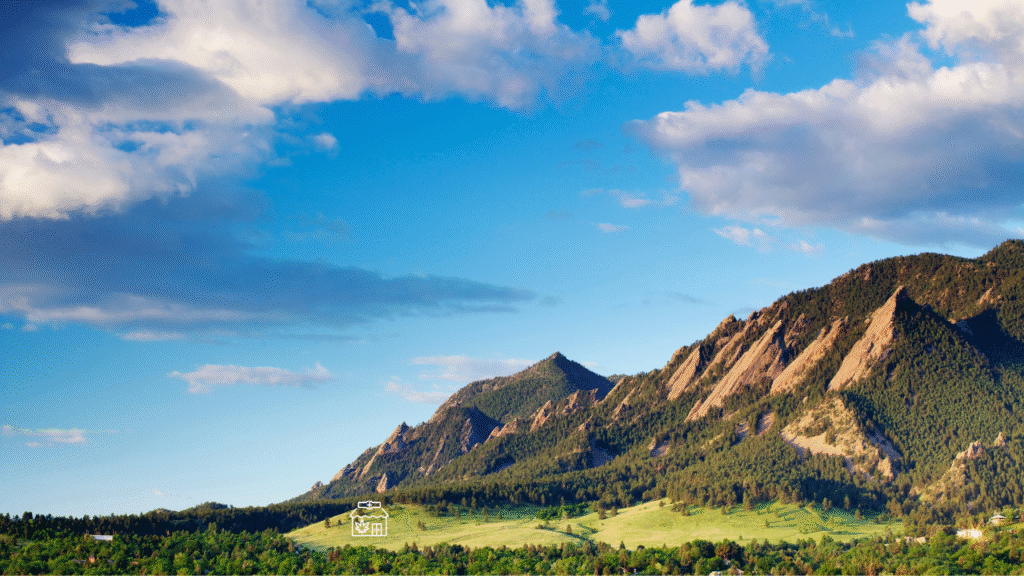 View of the Flatirons mountains near Louisville, Colorado, under a bright blue sky dotted with fluffy white clouds.