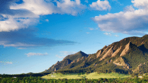View of the Flatirons mountains near Louisville, Colorado, under a bright blue sky dotted with fluffy white clouds.